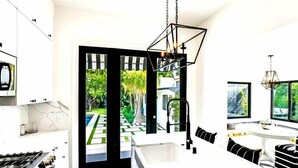 A modern kitchen with a central island, a dining area with a white table and black and white striped chairs, and a view of a garden through a glass door.