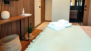 Hotelkamer met een houten paneelwand, een flatscreen televisie, een nachtkastje met een boek en een vaas, een netjes opgemaakt bed met handdoeken, een klein geweven mandje en een potplant.