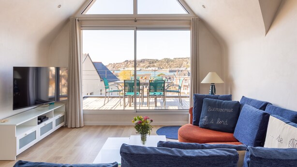 A modern living room with a large TV, comfortable seating, a view of a cityscape through a large window, and a balcony with outdoor furniture. - L'Abri Côtier d'Argäe - Terrasse vue mer (Erquy)