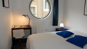A hotel room with a round window, a desk with a chair, a bed with blue pillows, and a bedside table with a lamp. - Three semi-detached houses downtown Grenoble (Grenoble)