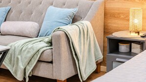 Un sofá gris con una manta verde claro, una almohada azul y otra almohada gris. Se observa una pared de madera y una mesita con una lámpara y un recipiente.