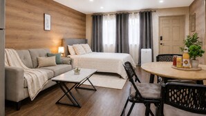 Una habitación de hotel moderna con cama, sofá, mesa de comedor, sillas, una mesita con una planta y un refrigerador.