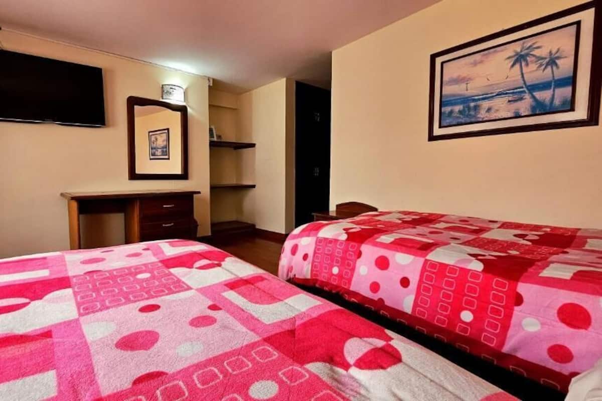 Ein Hotelzimmer mit zwei Betten, einem Schreibtisch, einem Stuhl, einem Fernseher und einem gerahmten Bild an der Wand.