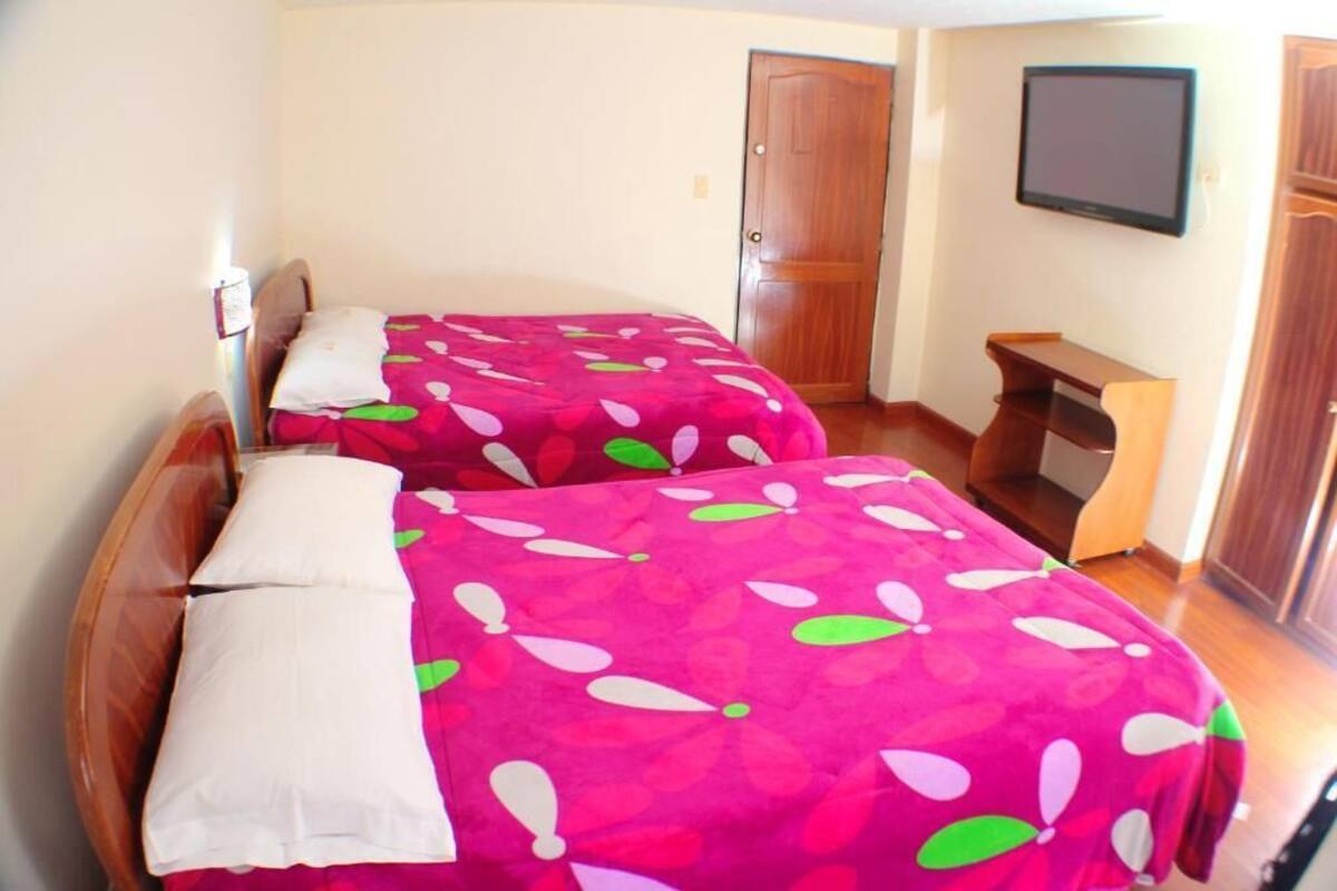 Ein Zimmer mit zwei Betten, jeweils mit einer zarten, blumigen Rosa-Decke, einer hölzernen Bank, einem an der Wand befestigten Fernseher und einer Holztür.