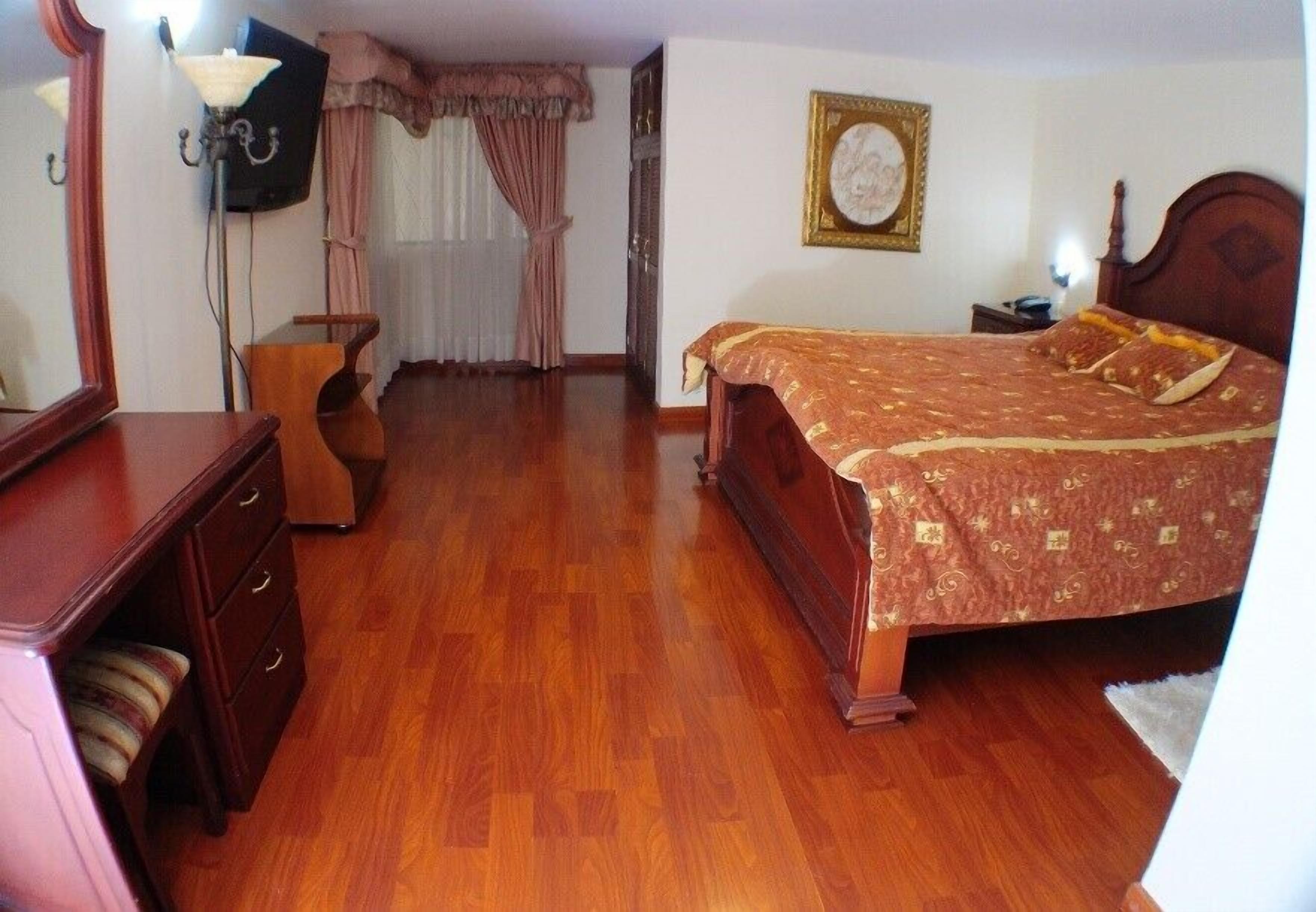 A bedroom with a wooden floor, a bed with a patterned bedspread, a dresser, a nightstand, a lamp, and a framed picture on the wall.