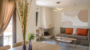 A modern living room with a fireplace, a sofa with orange cushions, a dining table, and a vase with tall plants. - Niriides Villas | Nymfe | Villa 2-bedrooms with private pool (Hersonissos)