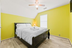 Un dormitorio con cama, ventilador de techo, ventana con persianas y mesita de noche con lámpara.