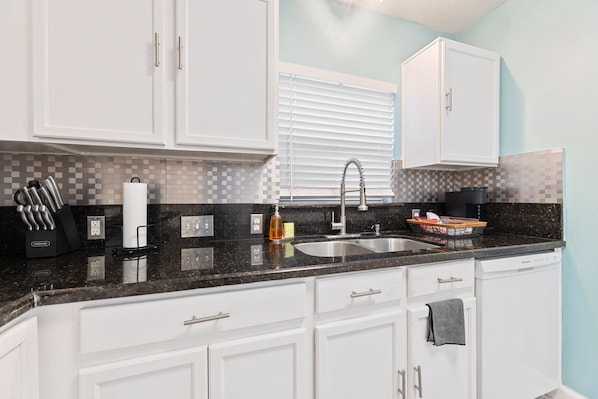 A modern kitchen with white cabinets, a granite countertop, a stainless steel sink, and a window with blinds.