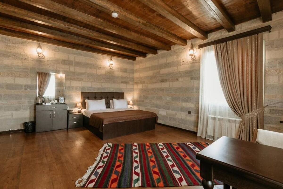 Una camera da letto con soffitto in legno, pareti in pietra, un letto con copriletto marrone, un comodino con lampada, una cassettiera e un tappeto decorato.