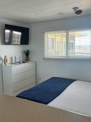 Un dormitorio con cama, cómoda, televisor, ventana con persianas y una planta.