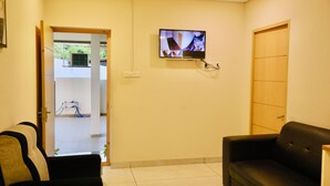 A room with a TV mounted on the wall, a sofa, and a door leading to another room. - Virksham Hotel (Coimbatore)
