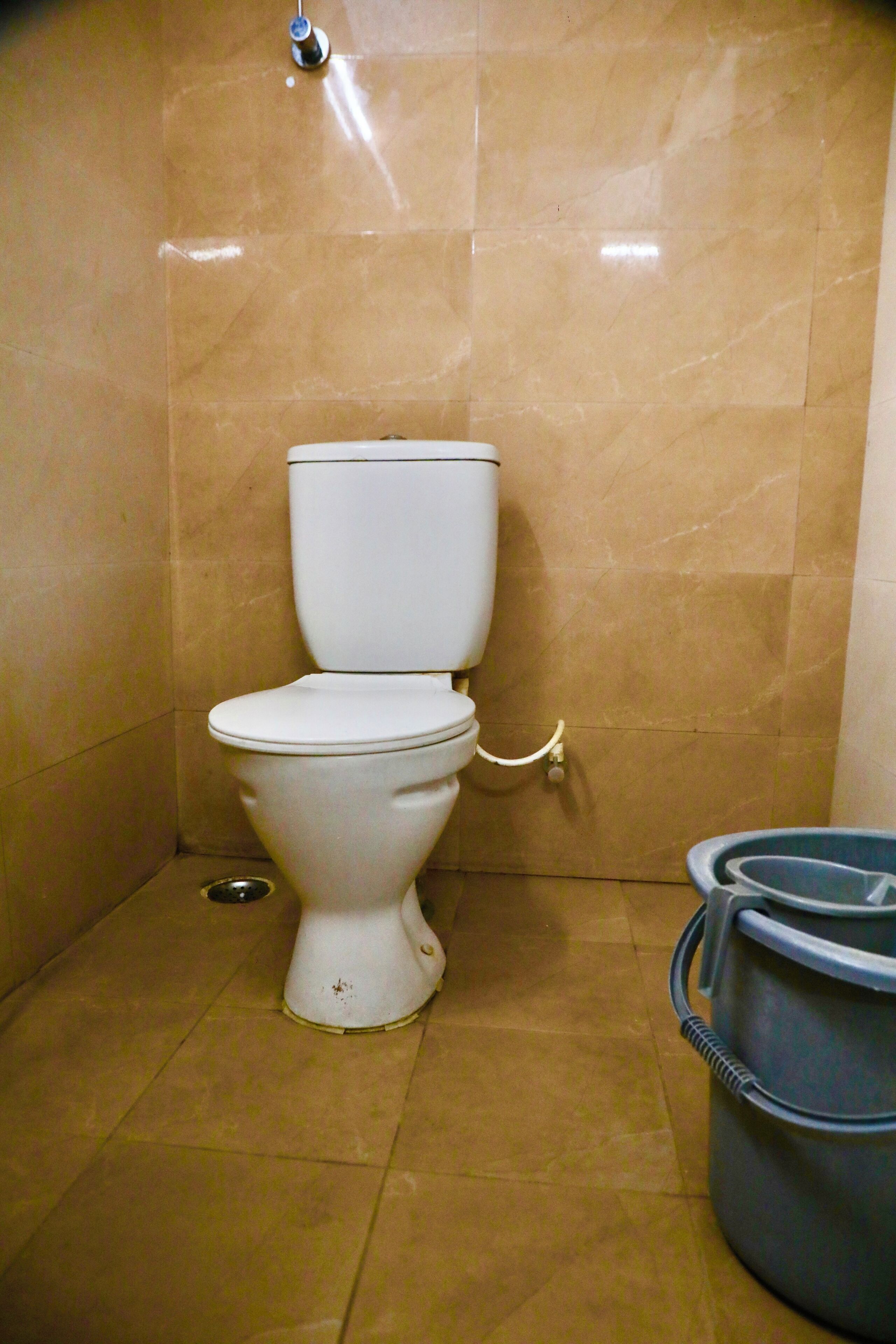 Ein kleines Badezimmer mit Toilette, einem Metallablauf und einem blauen Eimer.