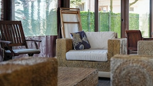 Een gezellige zithoek met een bank en stoelen, een houten stoel, en een groot raam met uitzicht op groen en een gebouw.