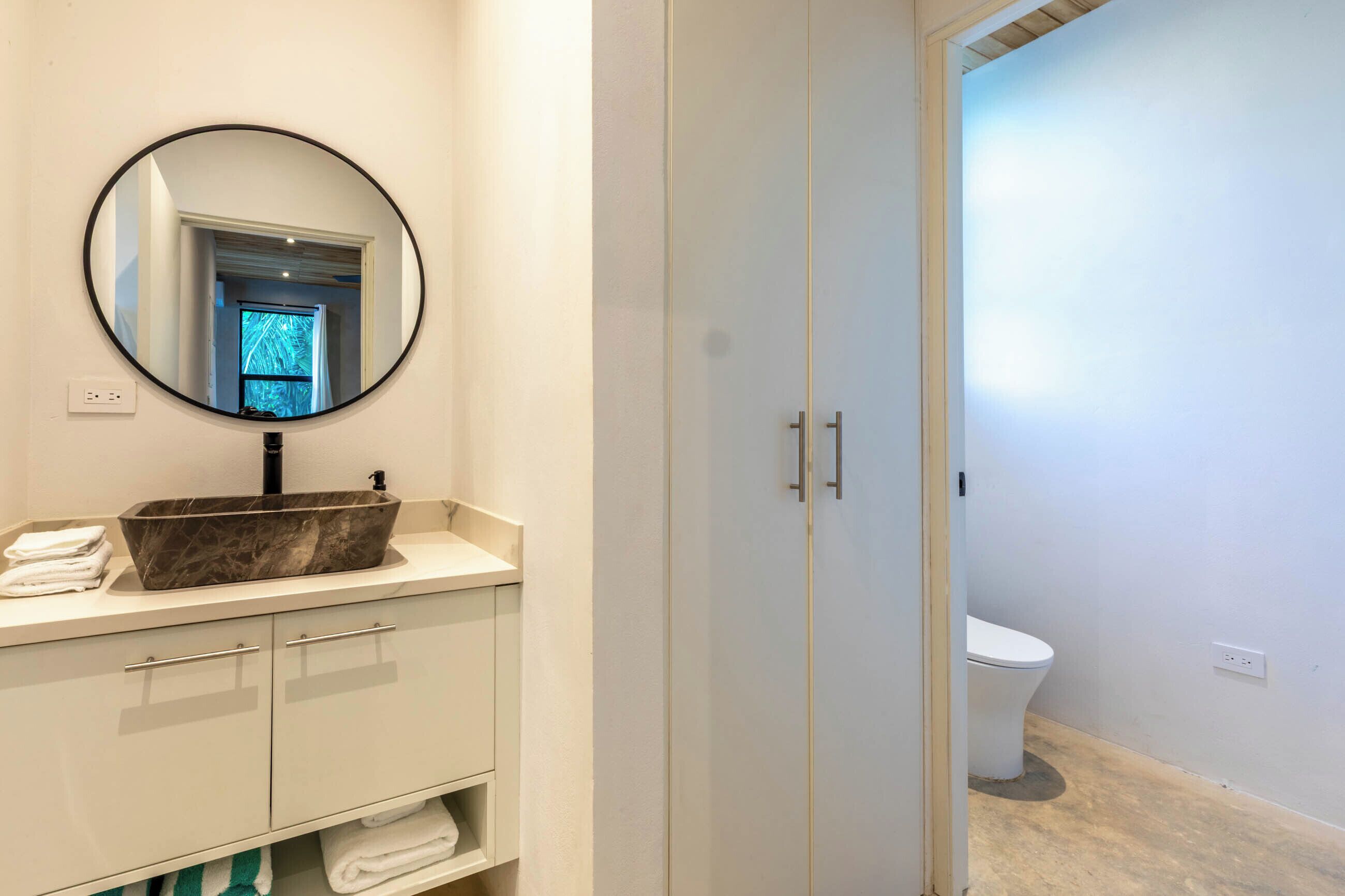 Une salle de bain avec un lavabo en marbre, un miroir rond, des toilettes et un placard.