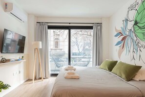 Ett hotellrum med en säng, en TV, ett skrivbord med en lampa, en stol och utsikt över staden genom stora fönster.
