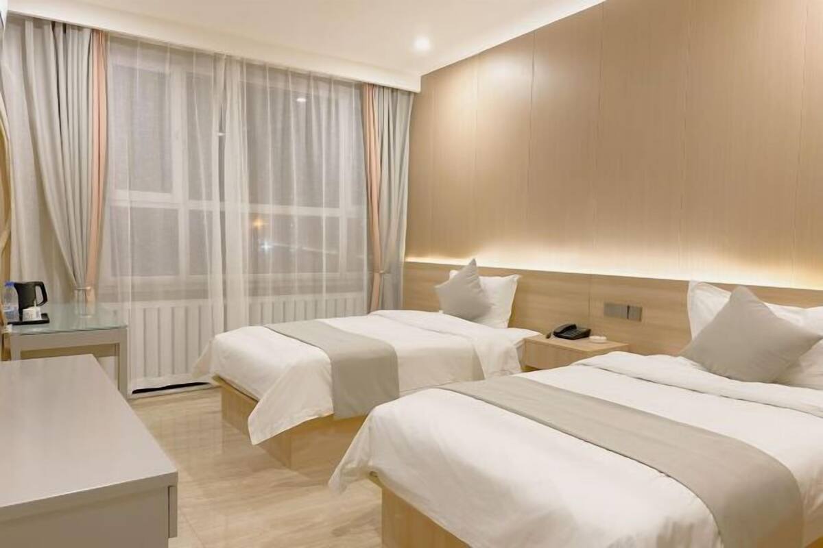 A hotel room with two beds, a bedside table, a lamp, and a view of the outside through sheer curtains.
