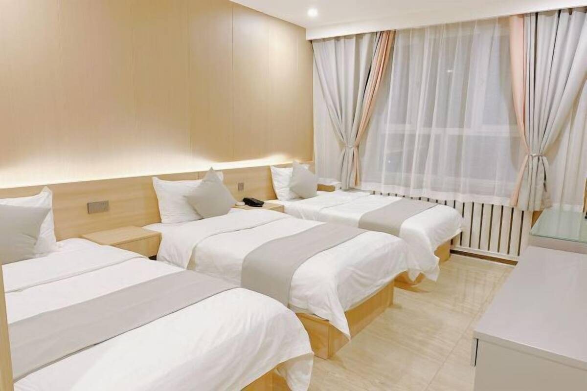 A hotel room with three beds, a bedside table with a lamp, a window with sheer curtains, and a white wall.
