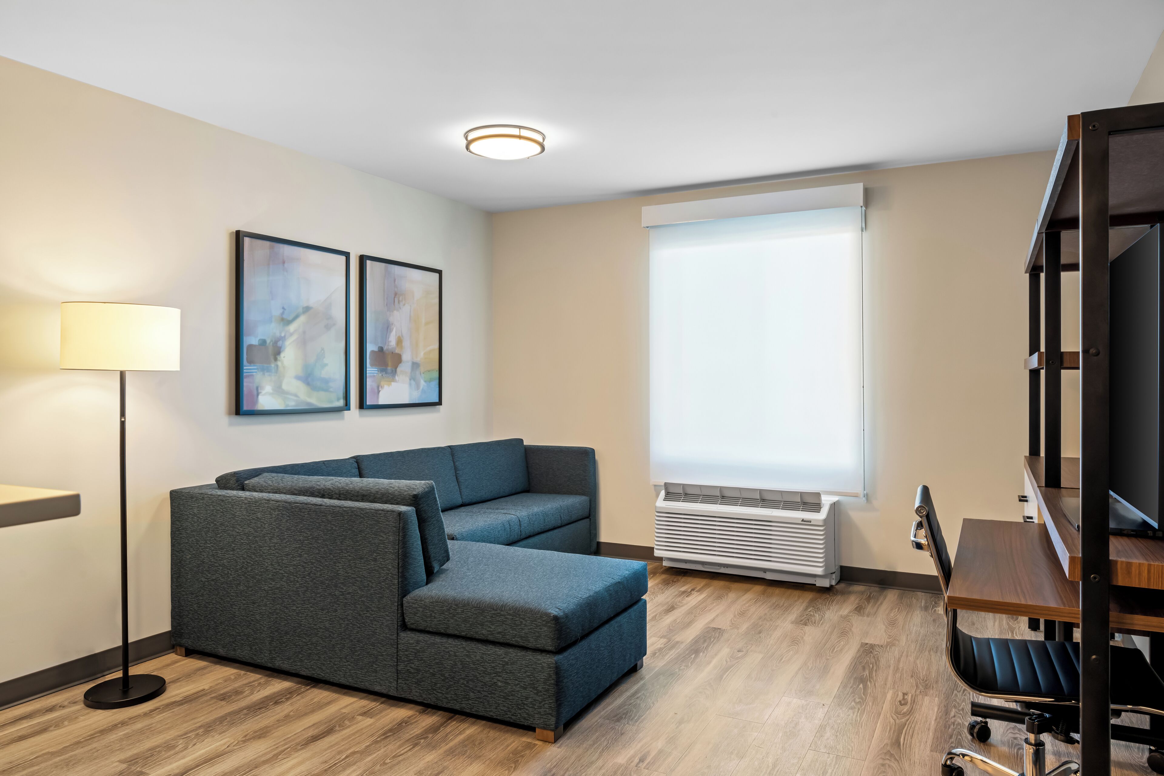 Un salon moderne avec un canapé d’angle, un lampadaire, un téléviseur à écran plat et une grande fenêtre équipée de stores.