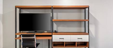 A modern office desk with a flat-screen monitor, a black office chair, and open shelving.