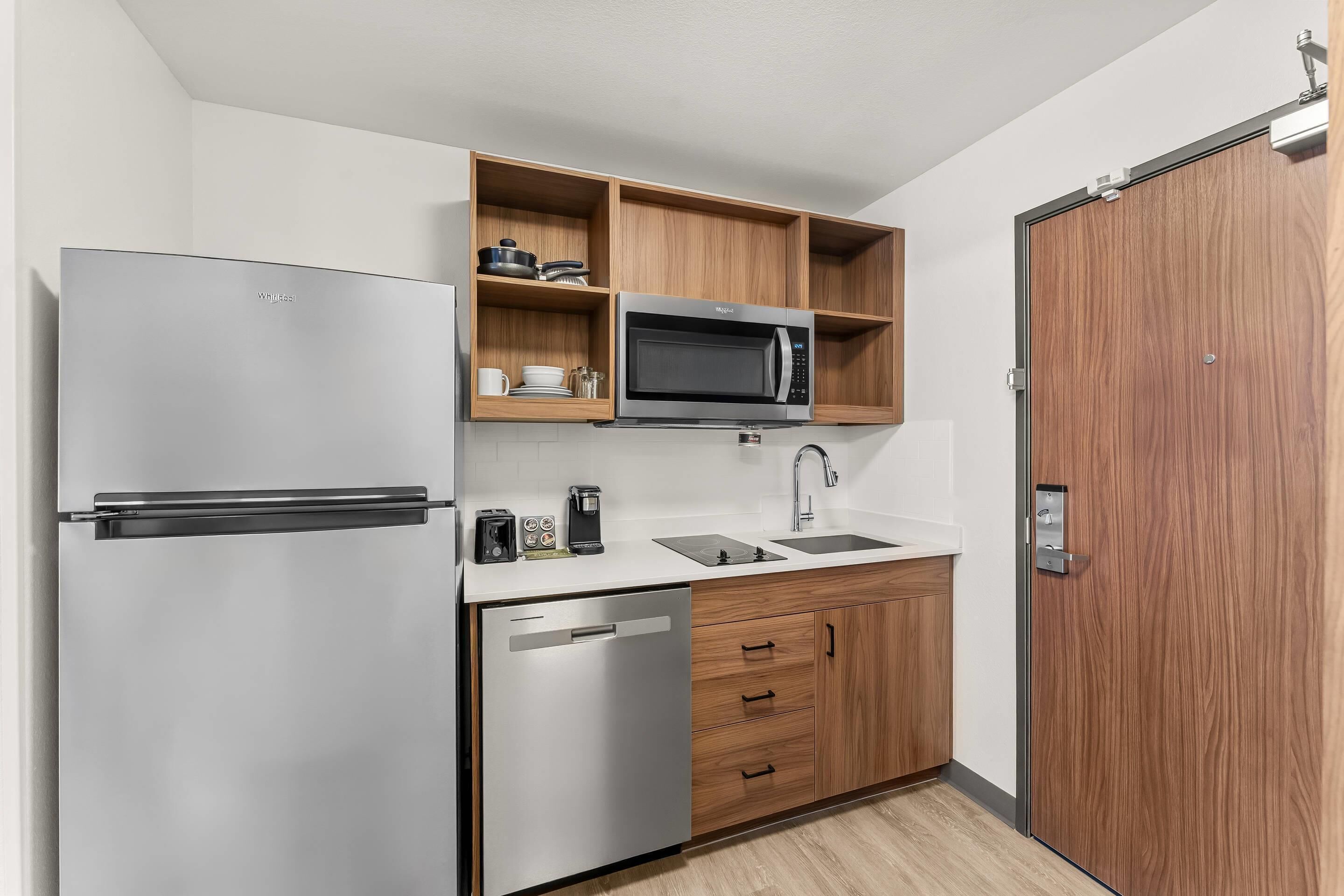 A compact kitchen with a stainless steel refrigerator, a microwave, a dishwasher, wooden cabinets, and a wooden door.