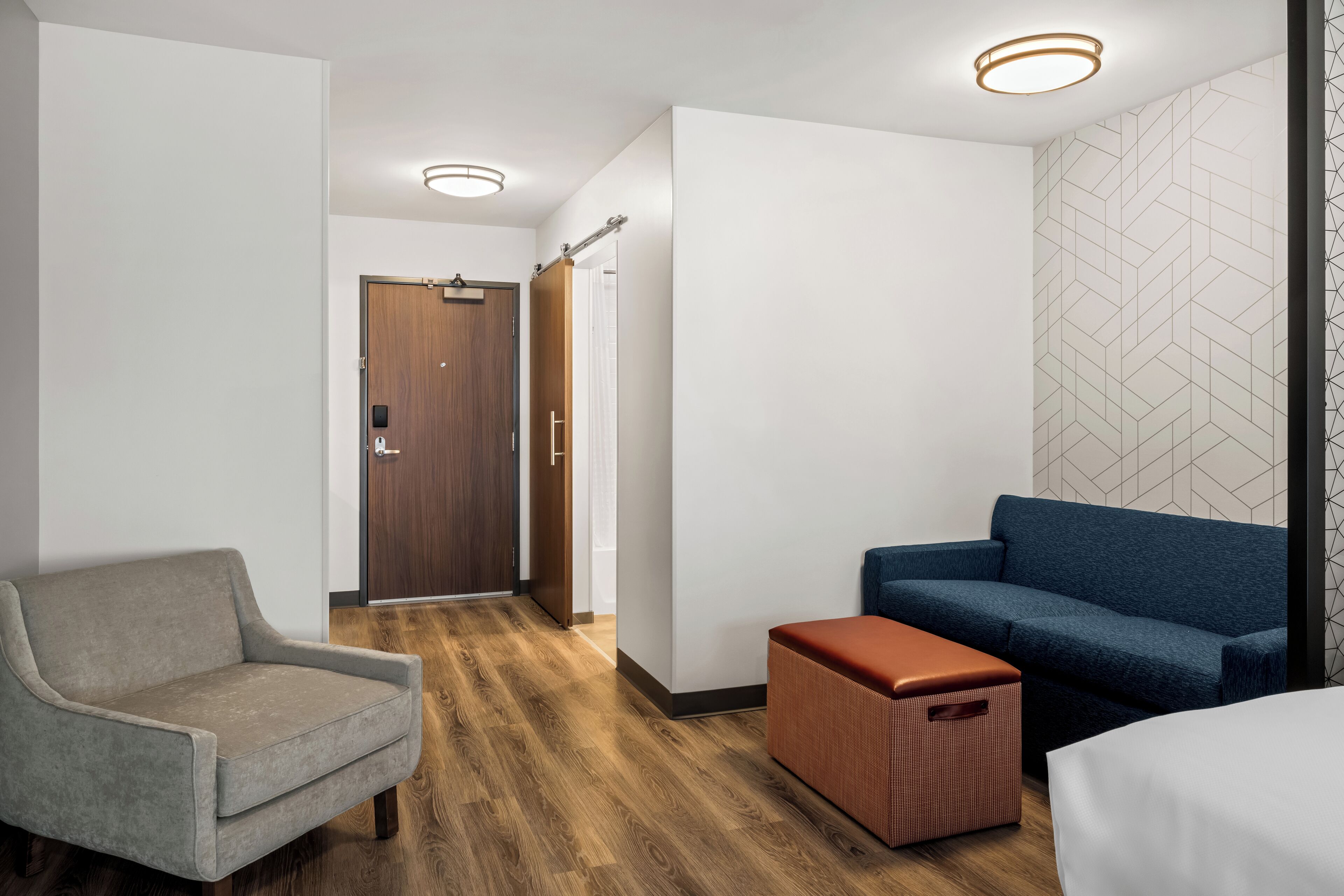 A modern hotel room with a wooden floor, a grey armchair, a blue sofa, and a small red ottoman.