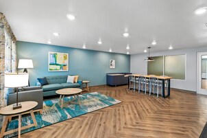 Una habitación de hotel moderna con una pared azul de acento, un sofá, una mesa de comedor con sillas, una lámpara y un ventanal grande.