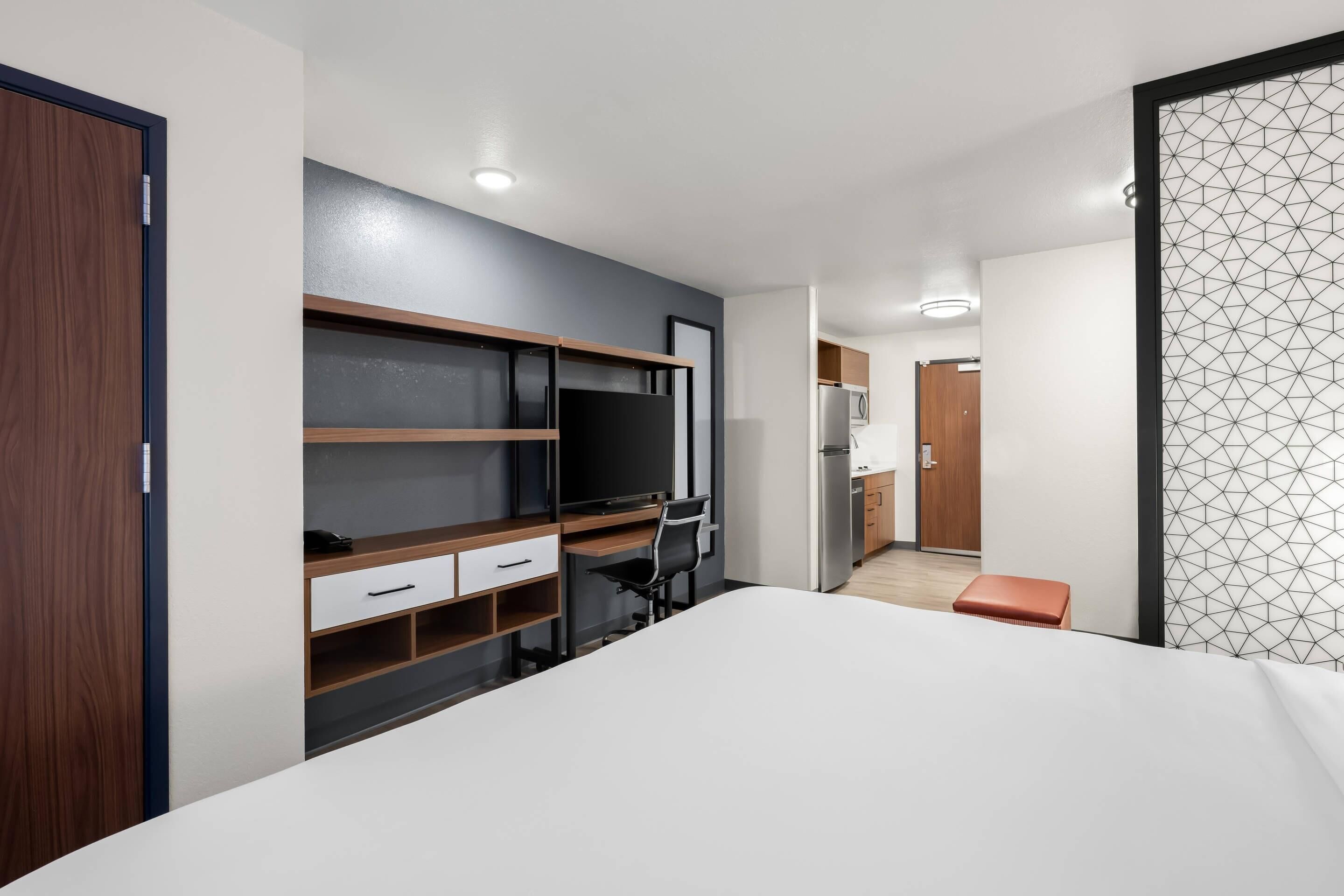A modern hotel room with a bed, a desk with a chair, a TV, and a kitchenette in the background.