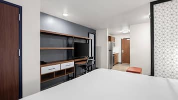 A modern hotel room with a bed, a desk with a chair, a TV, and a kitchenette in the background.