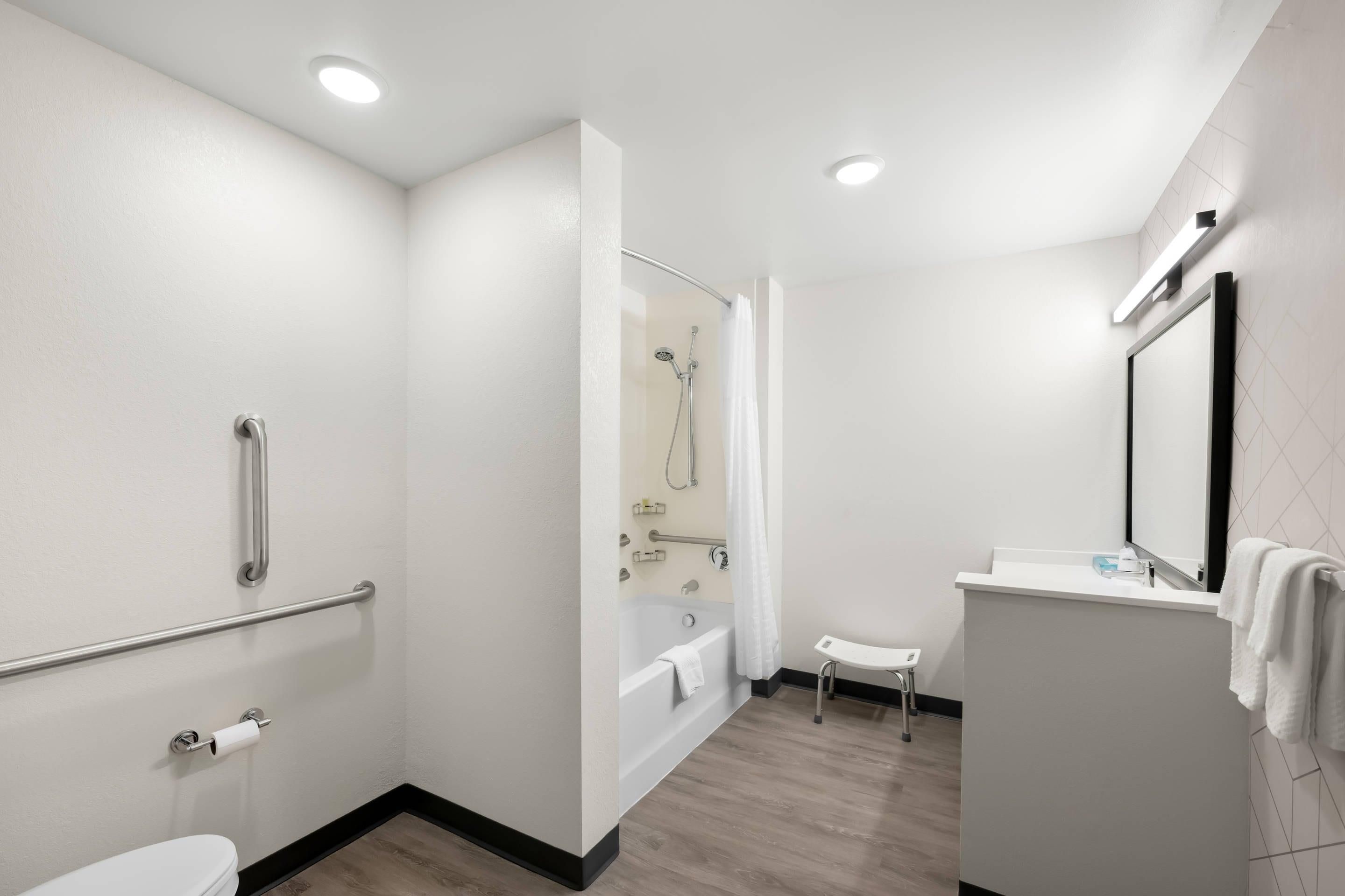 A modern bathroom with a bathtub, toilet, and sink, equipped with handrails and a shower area.