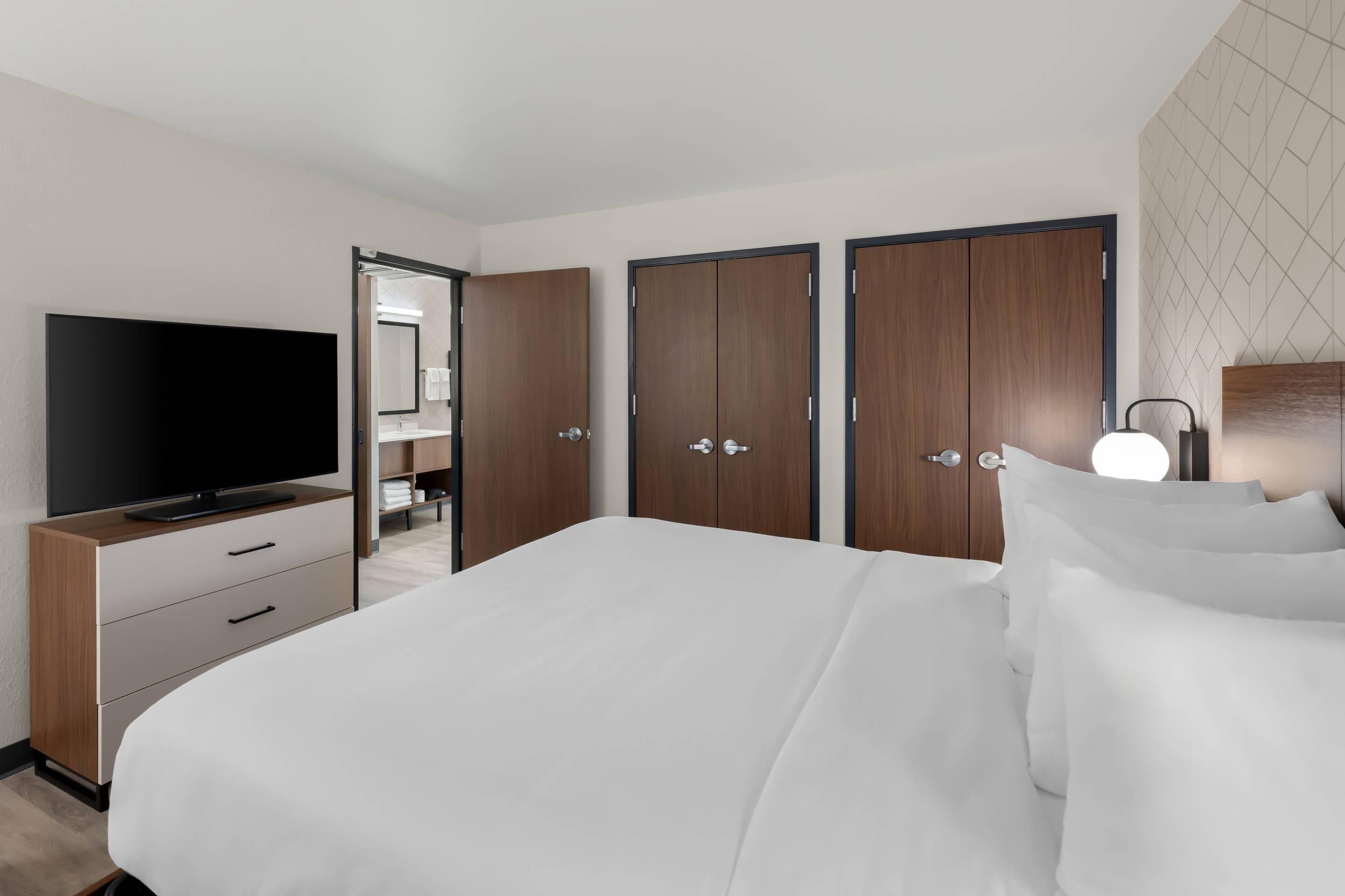 Habitación de hotel con una cama grande, televisión, armario y baño visible a través de una puerta abierta.