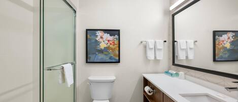 A modern bathroom with a glass shower enclosure, a white toilet, a wooden vanity with a white sink, and two framed abstract paintings on the walls.