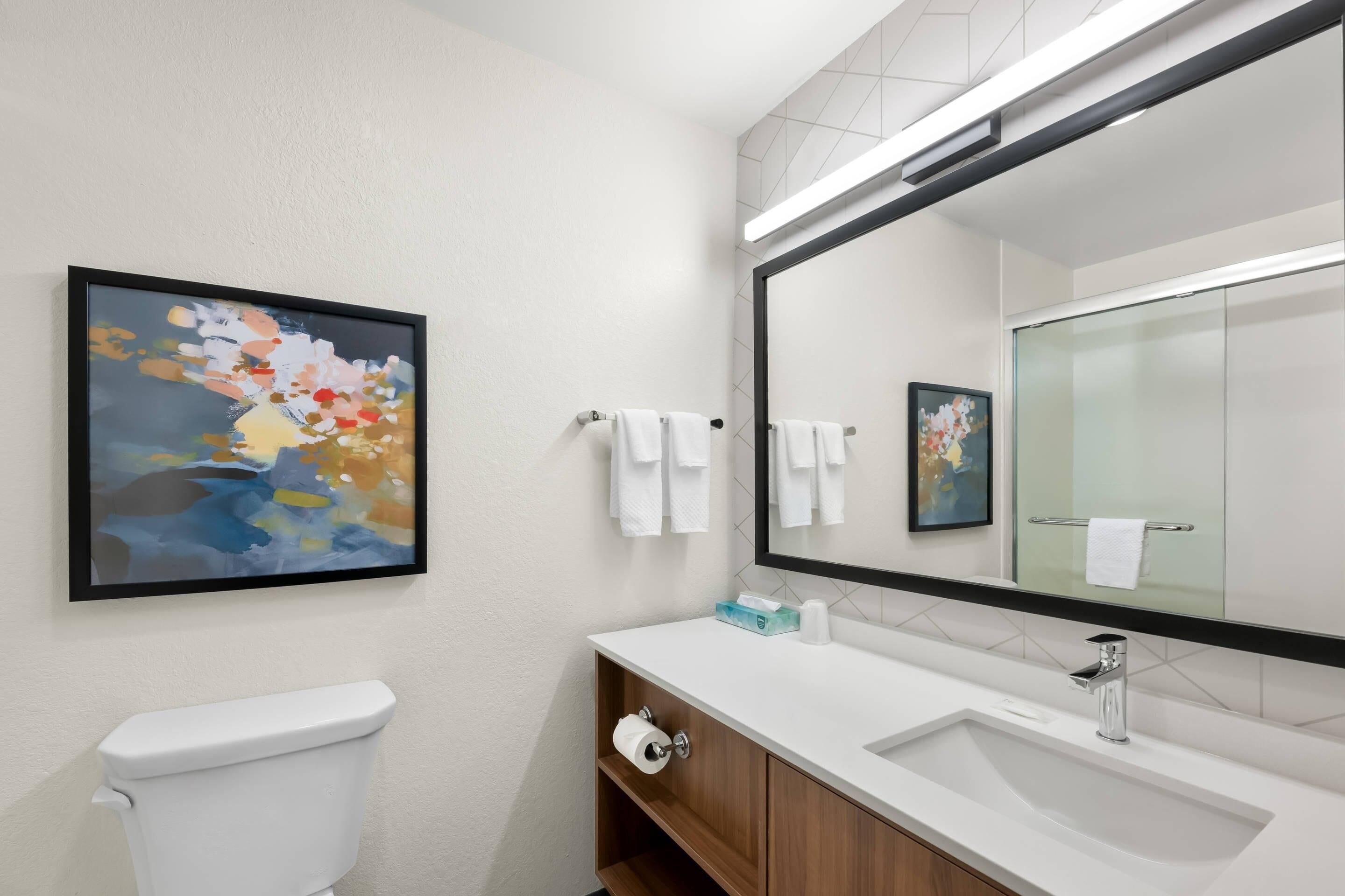 A modern bathroom with a toilet, sink, and mirror. There are towels hanging on the wall and a framed picture above the sink.