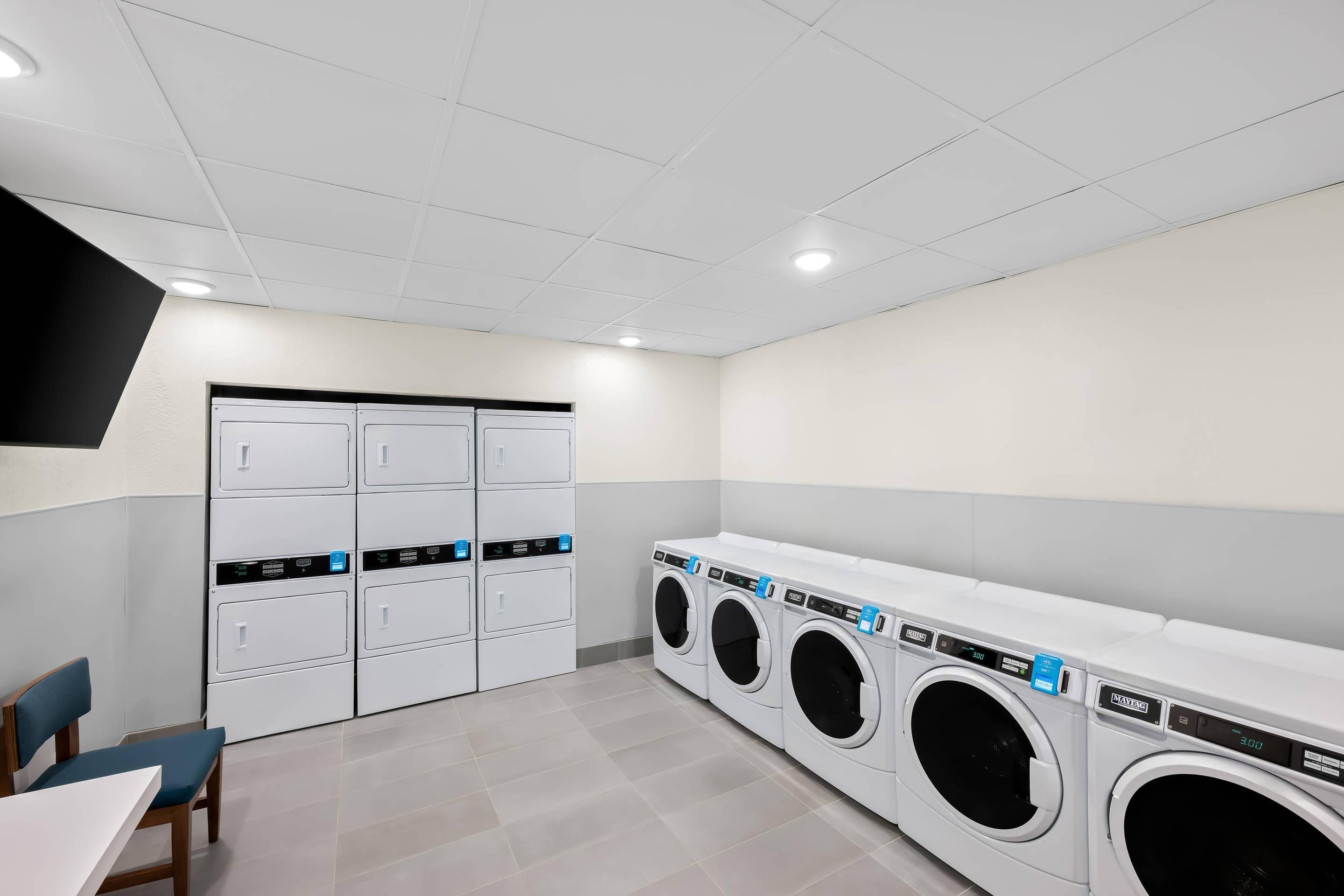 A laundry room with multiple washing machines and dryers, a flat-screen TV mounted on the wall, and a table with chairs.