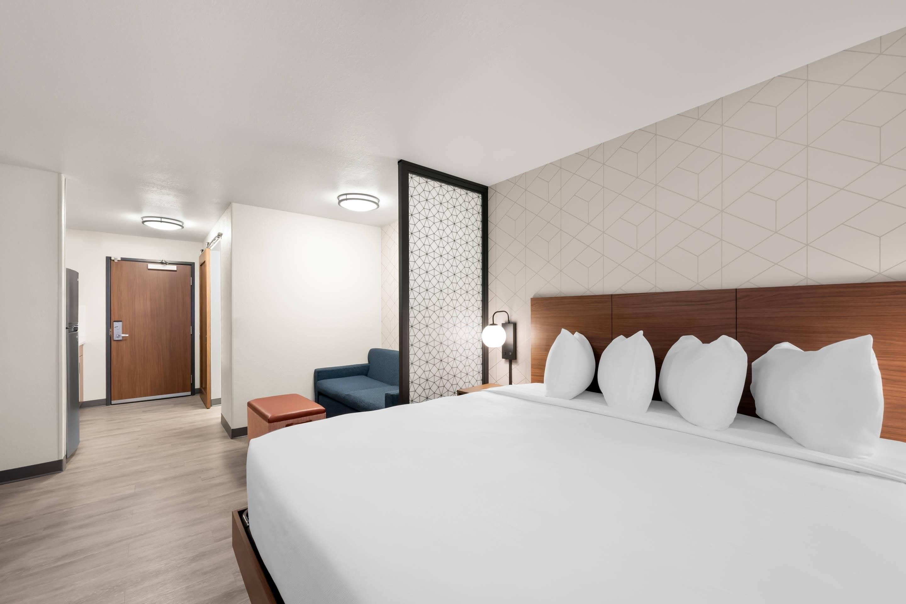 A hotel room with a large bed, a blue chair, a wooden headboard, and a geometric-patterned wall.