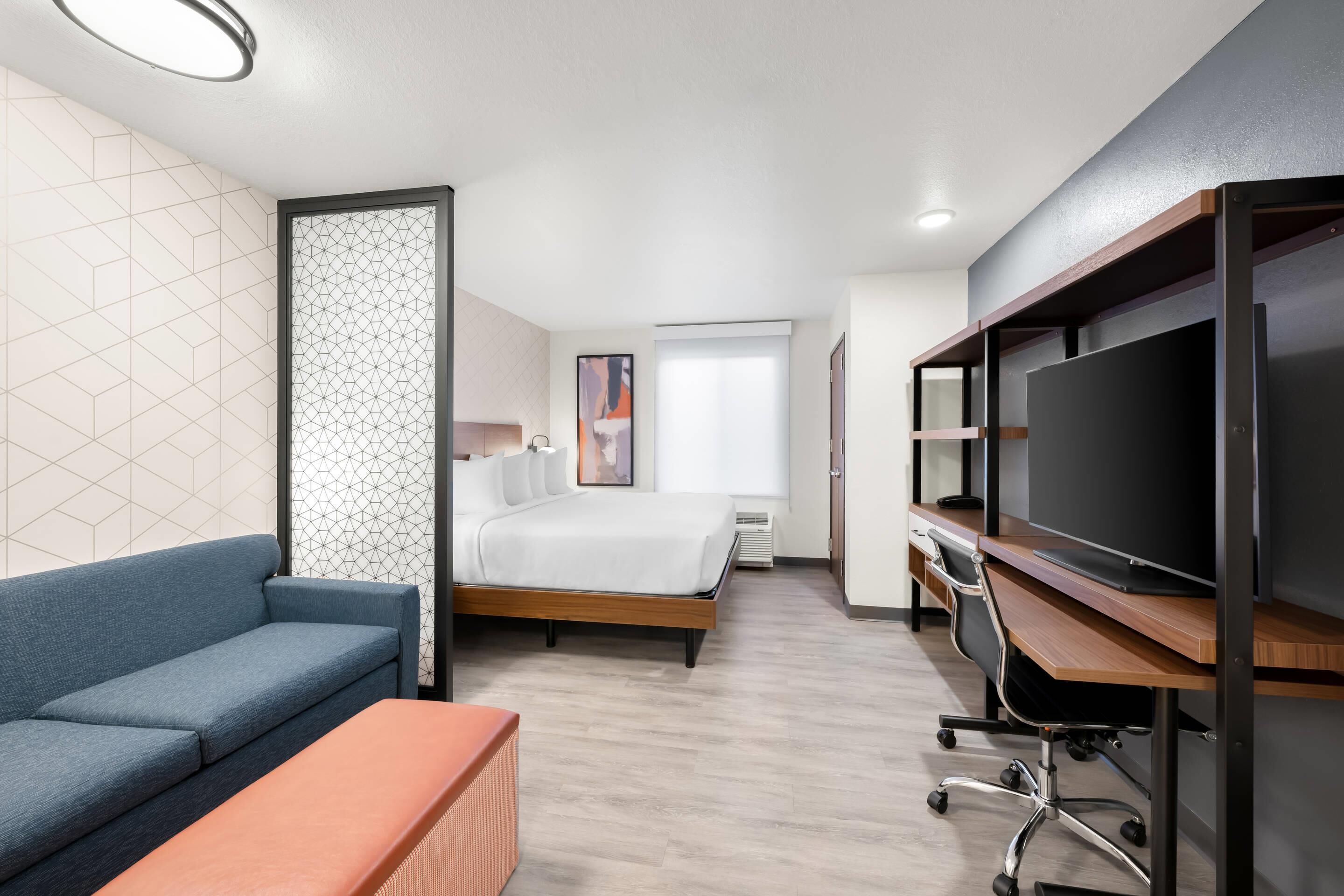 A modern hotel room with a bed, a sofa, a desk with a computer, and a TV.