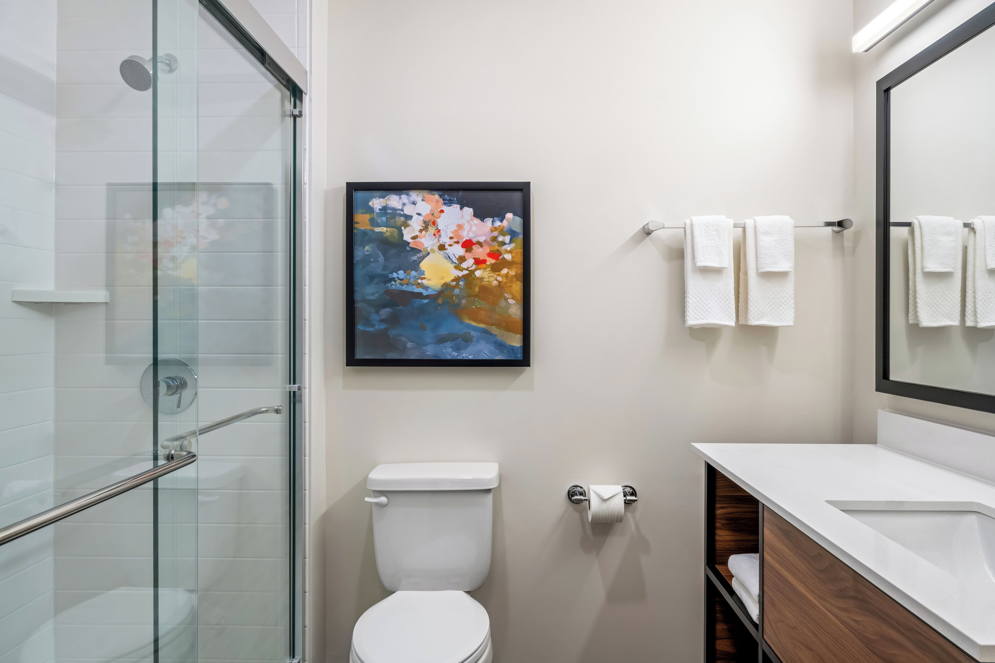 A modern bathroom with a glass shower enclosure, a white toilet, a framed abstract painting, and a vanity with a mirror.
