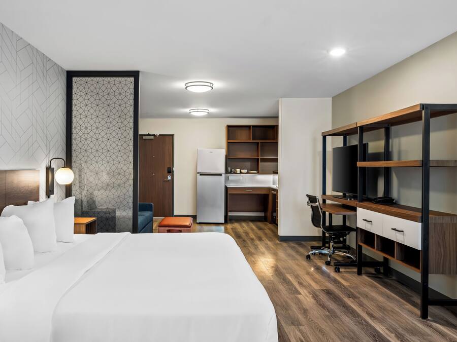 Une chambre d’hôtel avec un grand lit, un bureau avec un ordinateur, une télévision et, au fond, une petite cuisine.