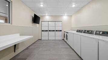A laundry room with a white sink, three washing machines, and a flat-screen TV on the wall.