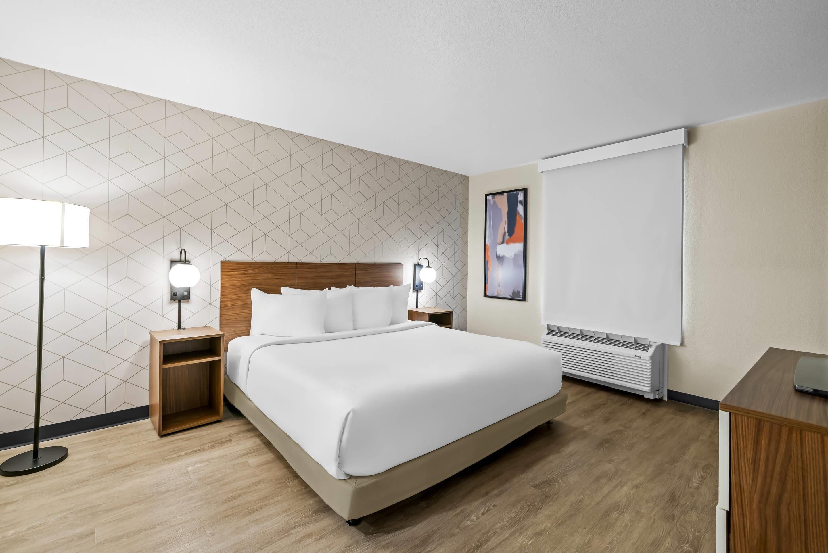 Habitación de hotel con cama, mesitas de noche, una lámpara de pie, aire acondicionado instalado en la pared y un cuadro.