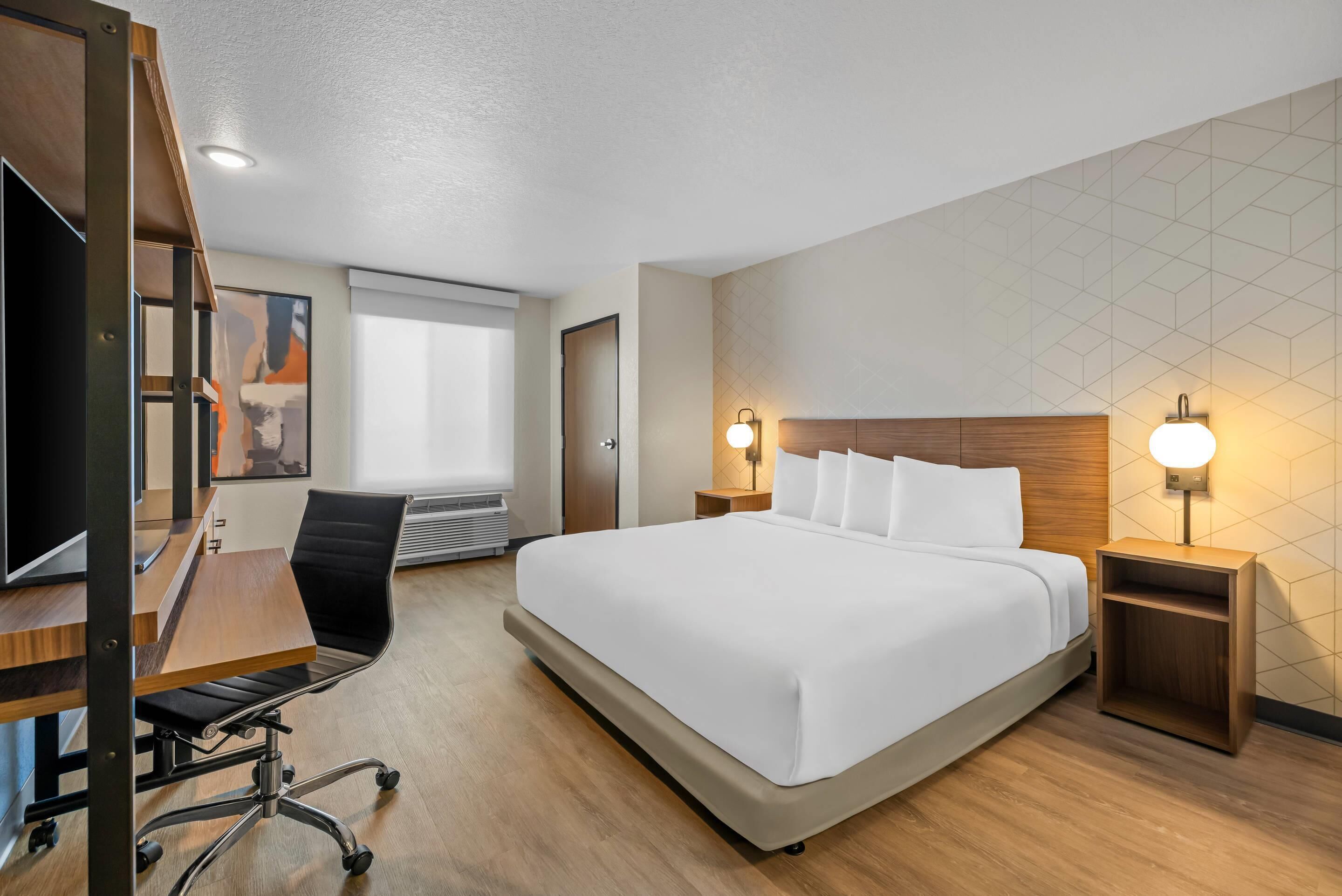 A hotel room with a large bed, a desk with a chair, a TV, and a window with blinds.