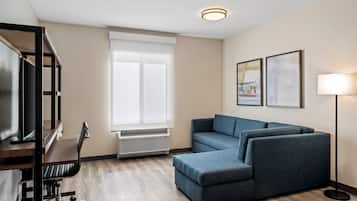 A modern hotel room with a wooden desk, a blue sofa, a standing lamp, and framed artwork on the wall.