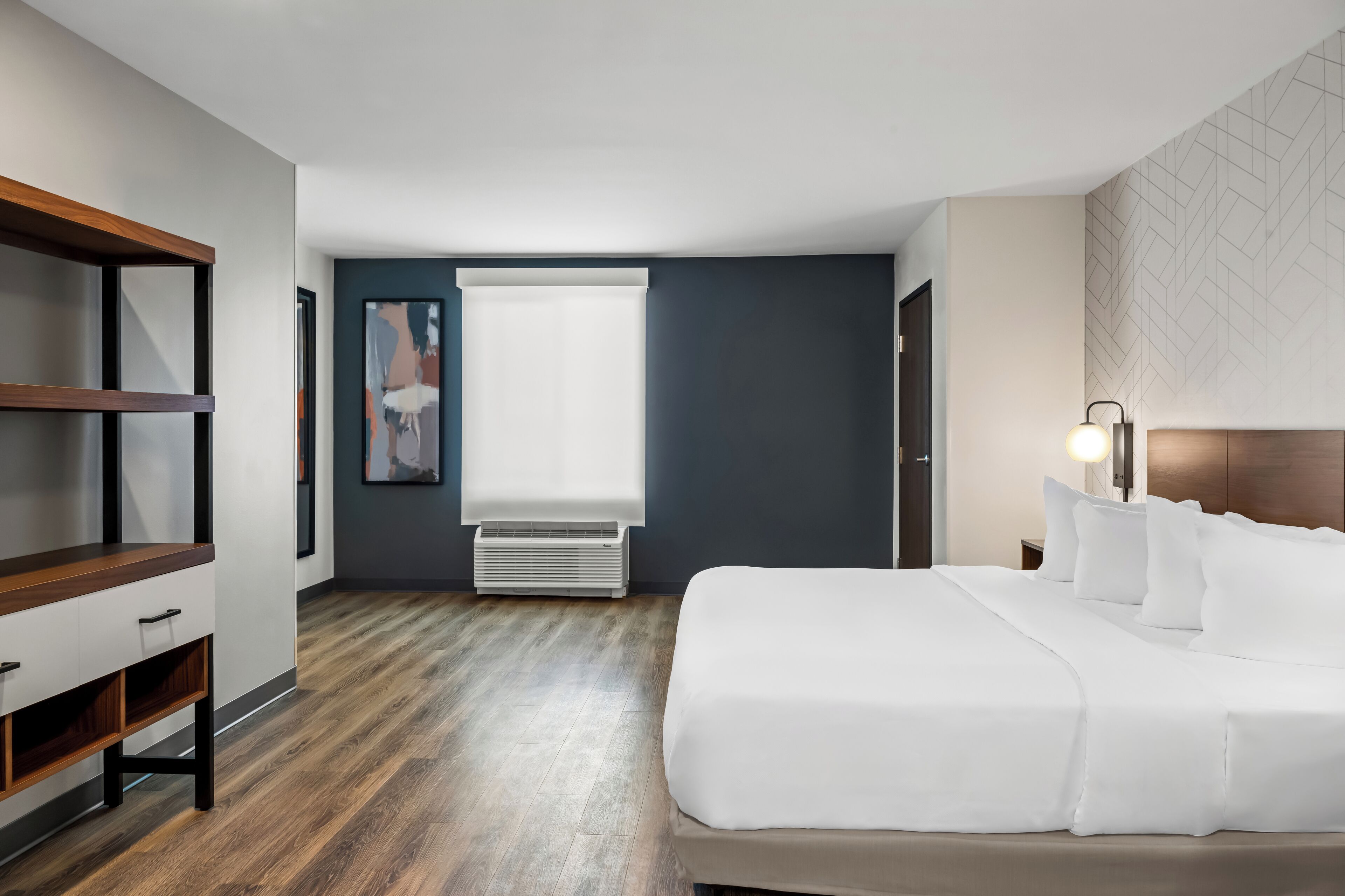 A modern hotel room with a large bed, a wooden nightstand, a desk, and a window with blinds.