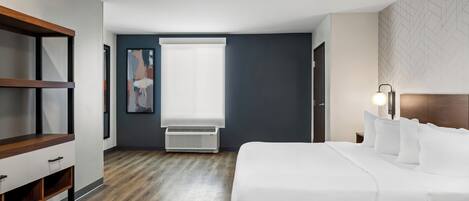 A modern hotel room with a large bed, a wooden nightstand, a desk, and a window with blinds.