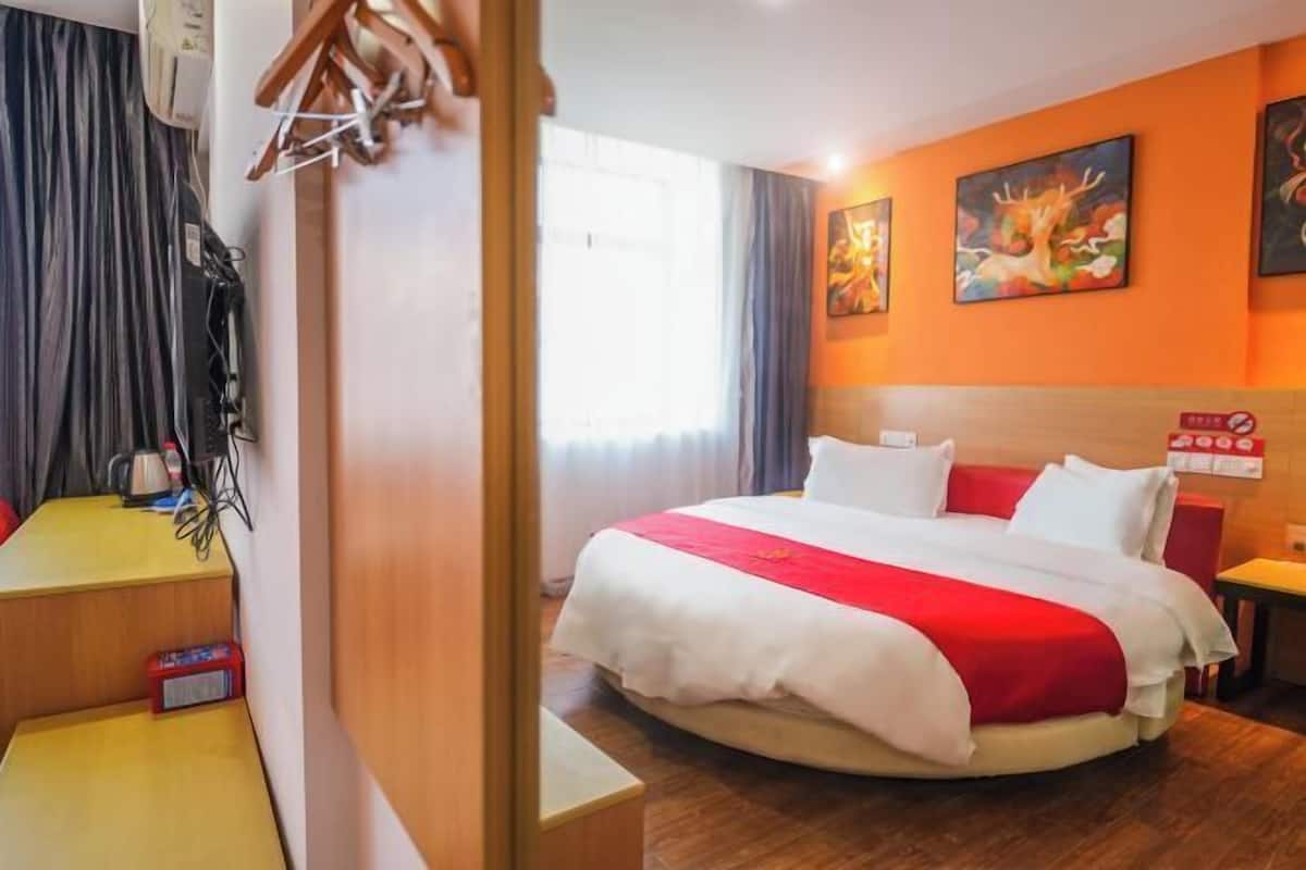 A hotel room with a bed, a desk, and a chair. There is a window with curtains, a painting on the wall, and a sign with Chinese characters.