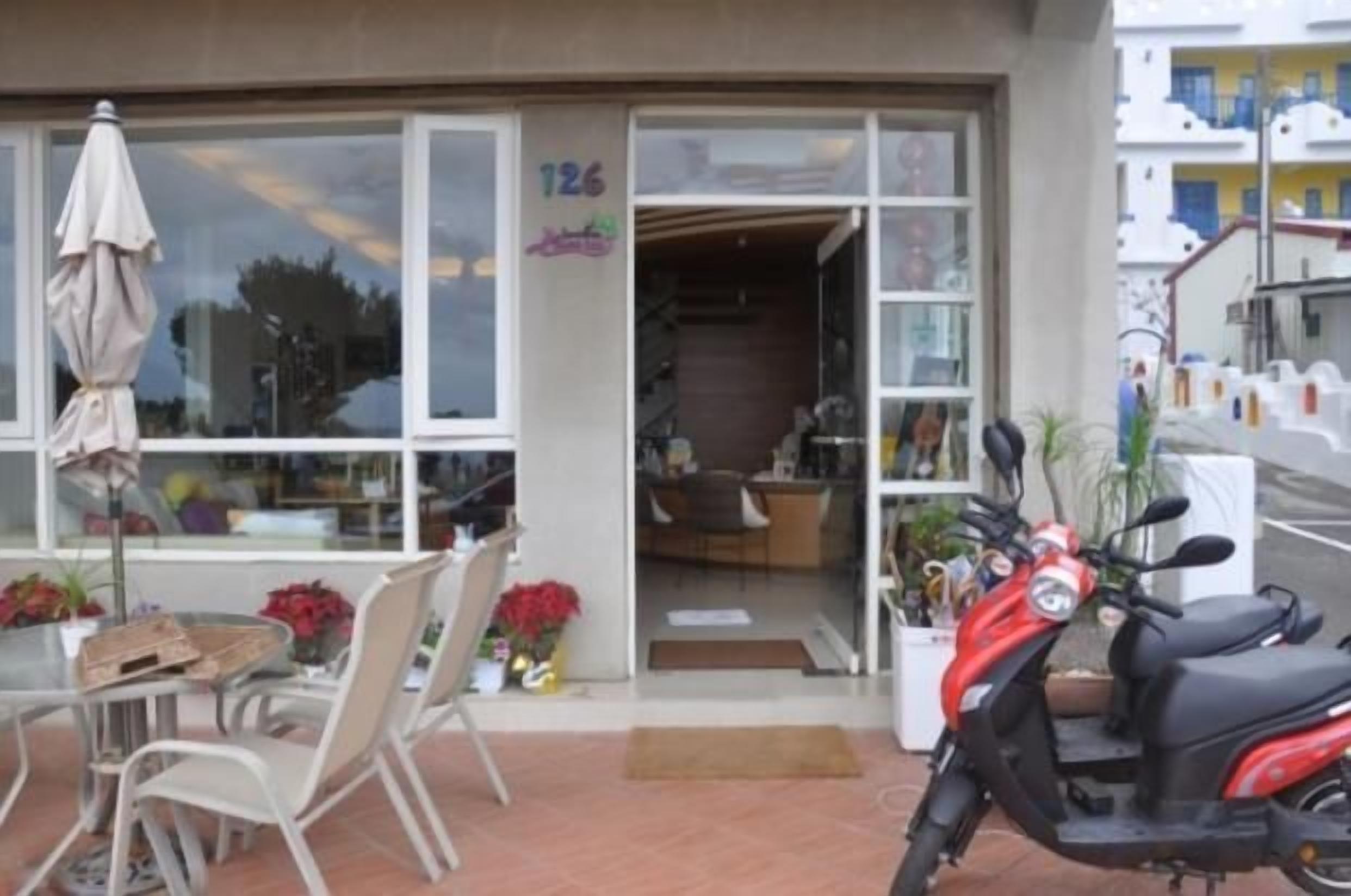 A storefront with a visible entrance, a scooter parked outside, and a small outdoor seating area with chairs and a table.