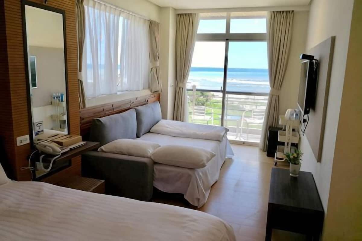 Habitación de hotel con dos camas, un escritorio, un espejo, una ventana con cortinas y vista al océano.