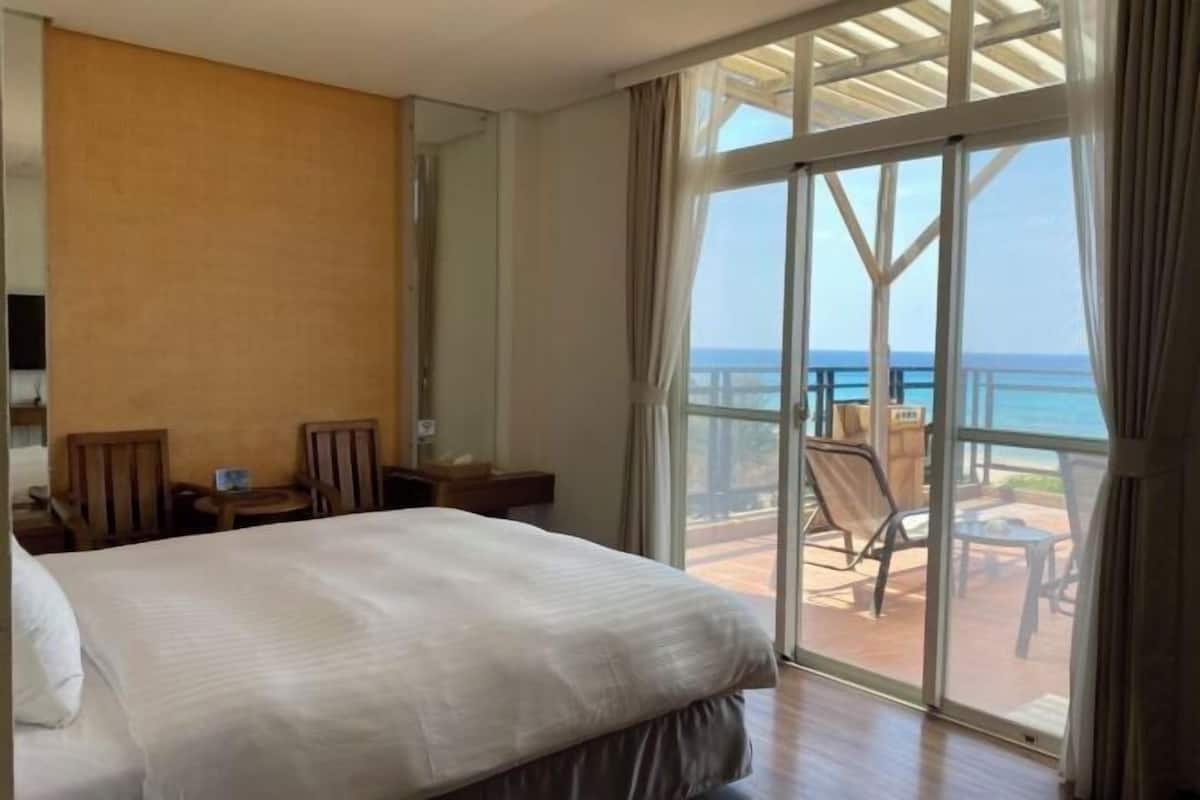 Habitación de hotel con una cama, un escritorio, una silla y vista al océano a través de amplias ventanas.