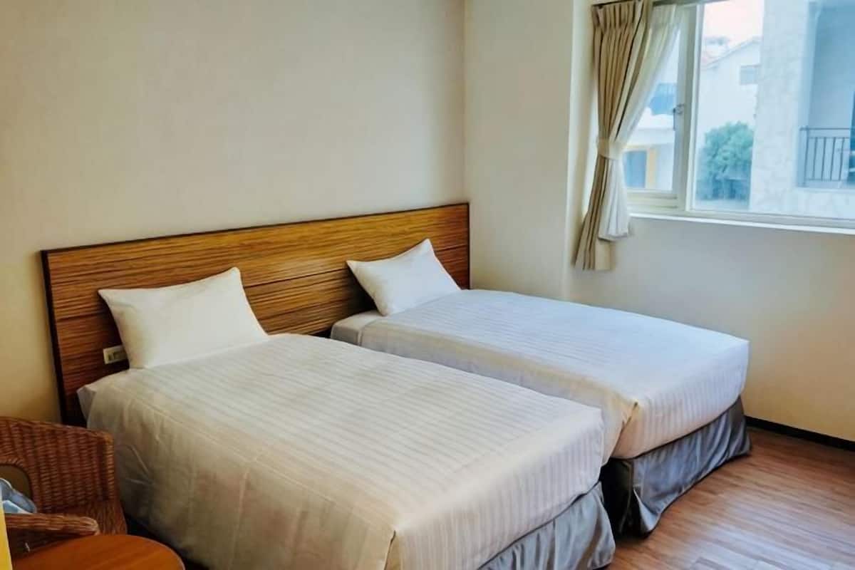 Dos camas individuales con cabeceros de madera, una silla de mimbre y una ventana con cortinas.