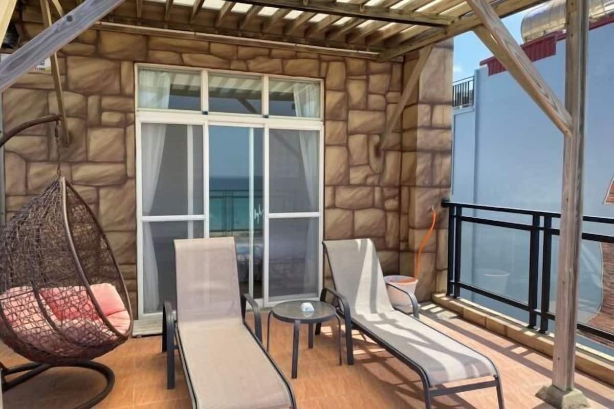 Un balcón con pared de piedra, una silla colgante, dos sillones, una mesita y una puerta de cristal que da a un espacio exterior.