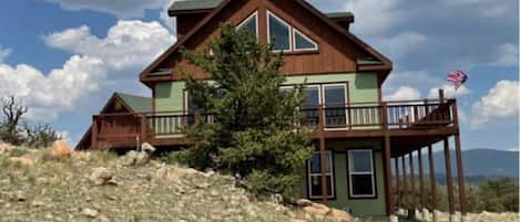 A two-story house with a green and brown exterior, a balcony with a railing, and an American flag on the right side.